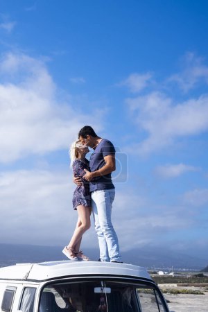 Photo for Love and travel people lifestyle - standing couple on the roof vehicle van together kissing and hugging with long road and blue sky in background for destination concept.- free lifestyle and alternative vacation - Royalty Free Image
