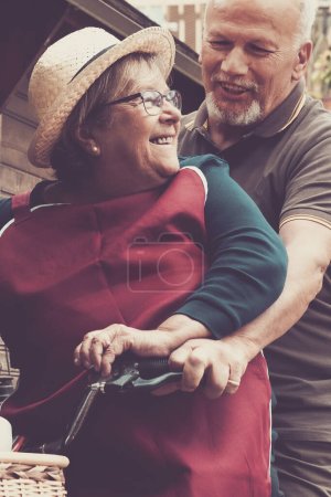Téléchargez les photos : Deux vieux hommes et femmes caucasiens s'amusent et aiment jouer ensemble en plein air avec un vélo et une maison en bois en arrière-plan heureux couple d'âge mûr sourire et rire - en image libre de droit