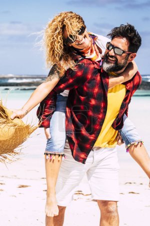 Téléchargez les photos : Le couple heureux profite de la relation et des vacances d'été ensemble en riant et en s'amusant - l'homme porte une femme et profite de l'activité de loisirs de plein air à la plage avec amour et amitié - en image libre de droit