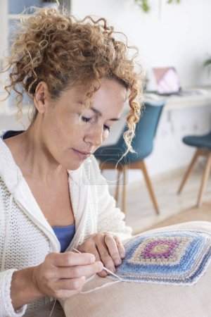 Photo for Adult caucasian woman at home doing crochet hobby activity at home decorating a shabby chic white pillow. Living room interior in background. Female people creating woolen decorations - Royalty Free Image