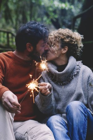 Téléchargez les photos : Couple adulte romantique embrassant en amour tenant des étincelles de feu assis dans le parc naturel profiter de relations en plein air et de loisirs ensemble. Concept de personnes heureuses profitant de vacances à la montagne - en image libre de droit
