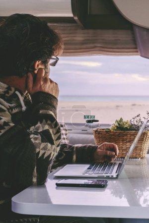 Téléchargez les photos : Man working inside camper van sitting at the table and looking outside the window the beautiful beach and ocean in background. Concept of freedom lifestyle and remote online worker people - en image libre de droit