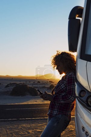 Foto de People on travel with modern camper. Woman using mobile phone roaming against big motorhome. Concept of modern lifestyle and travelers. Vacation and vanlife sending message with cellular. Everywhere internet connection - Imagen libre de derechos