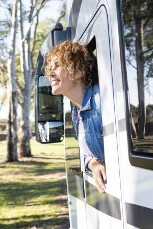 Foto de Hermosa mujer rizada caucásica sonriente inclinada por la ventana de una caravana. Viajes, naturaleza y ocio - Imagen libre de derechos
