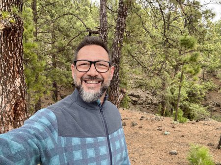 Photo for Adult man taking selfie picture with forest park woods in background. Mature using mobile phone to take picture during weekend activity wearing glasses and enjoying outdoor leisure activity travel - Royalty Free Image