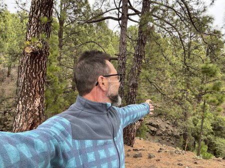 Téléchargez les photos : Homme adulte prenant des photos selfie avec forêt parc bois en arrière-plan. Mature utilisant un téléphone mobile pour prendre des photos pendant l'activité de week-end en portant des lunettes et en appréciant les loisirs de plein air - en image libre de droit