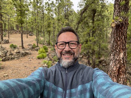 Téléchargez les photos : Heureux homme gai seul dans les bois prendre des photos selfie dans les loisirs en plein air seul avec des arbres et la forêt en arrière-plan. Voyageur moderne profitant d'une destination pittoresque à la montagne. Nature - en image libre de droit