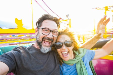 Photo for Adult couple have fun together on a roller coaster in amusement park taking selfie picture with the phone. People enjoying holiday lifestyle. Mature man and woman enjoy time outdoor leisure activity - Royalty Free Image