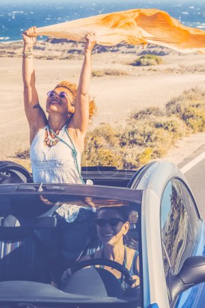 Photo for Car travel friends transportation two happy women enjoy convertible auto together in summer trip holiday vacation - ocean and beach in background - driving and having fun concept - Royalty Free Image