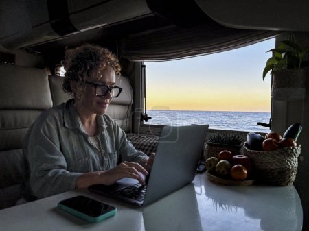 Photo for One modern adult woman digital nomad working on laptop sitting inside a motorhome camper van and using laptop computer with roaming traveler connection with phone on the table, Alternative job people - Royalty Free Image