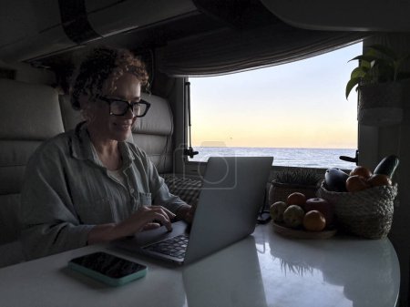 Téléchargez les photos : Femme adulte moderne nomade numérique travaillant sur ordinateur portable assis à l'intérieur d'un camping-car camping-car et en utilisant un ordinateur portable avec connexion itinérante voyageur avec téléphone sur la table, les gens de travail alternatif - en image libre de droit