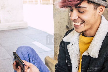 Téléchargez les photos : Jeune adolescent moderne assis sur le sol en utilisant l'application téléphonique pour le message et la vie sur les médias sociaux. en ligne connecté jeune garçon mâle avec des vêtements alternatifs de mode à la mode dans la vie urbaine fond - en image libre de droit