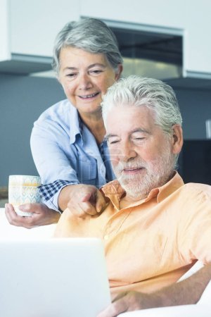 Photo for Cute senior man and woman using computer at home. Man sitting on the sofa and woman behind him watching the laptop display. Mature people surfing the web indoor. Elderly real lifestyle - Royalty Free Image