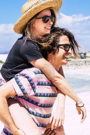 Foto de Joven pareja milenaria disfrutar de vacaciones en la playa en un destino turístico isla tropical. Chico y chica se divierten en la actividad de ocio al aire libre, llevándola a cuestas y riendo mucho juntos - Imagen libre de derechos