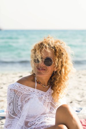 Foto de Alegre sonrisa turística femenina y disfrutar de vacaciones de verano sentado y relajante en la playa con agua de mar azul en el fondo. Arena tropical y mujer estilo de vida feliz. - Imagen libre de derechos