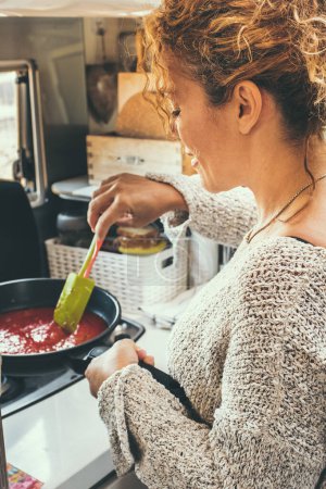 Téléchargez les photos : Femme vue de dos préparer et cuire la sauce tomate pour le déjeuner à l'intérieur d'un camping-car moderne. Mode de vie hors réseau et van avec les voyageurs ayant une préparation de nourriture. Activité de cuisine intérieure - en image libre de droit