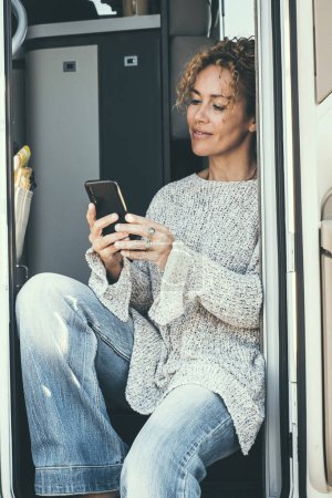 Photo for An independent adult woman sitting on the door of her modern motor home camper van use mobile phone connection and smile. Cheerful female with travel and technology leisure activity - Royalty Free Image