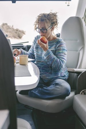 Photo for View of  woman sitting and having leisure time inside a camper van motor home. Healthy lifestyle female eating apple and using computer laptop. Alternative free off grid lifestyle - Royalty Free Image