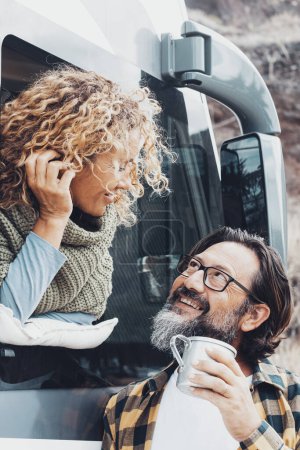 Photo for Man and woman smiling and using camper van transport vehicle. Couple enjoying vacation and leisure. Female outside the motor home window speak with male drinking coffee. Freedom off grid life - Royalty Free Image