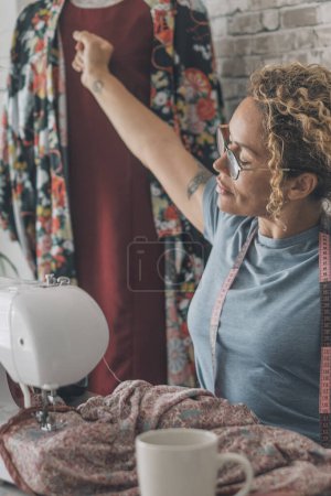 Foto de Mujer extendiendo los brazos después del trabajo en la máquina de coser en casa o estudio de diseño de ropa. Cuerpo de estiramiento femenino después del exceso de trabajo con reparación o producción de ropa. Concepto de trabajo alternativo en interiores - Imagen libre de derechos