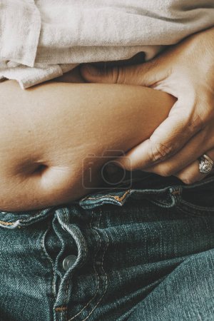 Photo for Close up of female holding her belly with fat. Unhappy body positivity concept. Woman thinking about diet. Lazy lifestyle. Eating disorder and unhealthy people. Overeating and feed. Changes baby born - Royalty Free Image