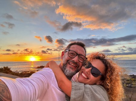 Foto de Romantic couple of man and woman in love taking selfie picture together hugging and smiling at the phone camera and enjoying beautiful sunset on the ocean at the beach. Travel people summer vacation - Imagen libre de derechos