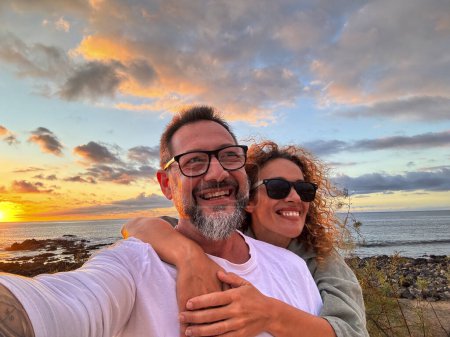 Photo for Happy adult couple in love taking selfie picture with romantic wonderful sunset on the ocean in background. Young mature people man and woman enjoying travel destination in summer holiday vacation - Royalty Free Image