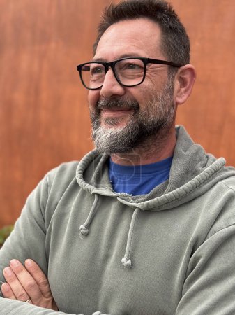Téléchargez les photos : Portrait d'un homme adulte souriant et regardant devant portant des lunettes et un pull vert sur un fond de mur brun dans les loisirs de plein air seul - en image libre de droit