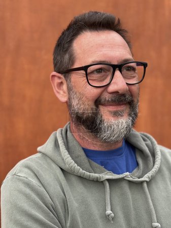 Téléchargez les photos : Portrait d'un homme adulte souriant et regardant devant portant des lunettes et un pull vert sur un fond de mur brun dans les loisirs de plein air seul - en image libre de droit