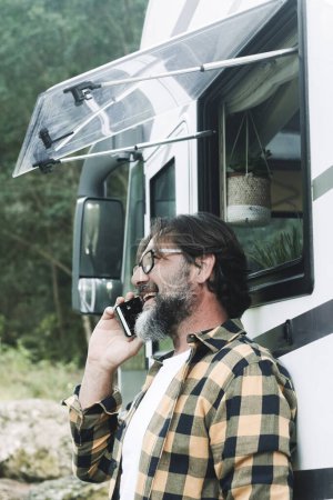 Photo for Man outside a motorhome do a phone call with a smile. People and alternative travel van life lifestyle. Alone adult male using phone. Nature park woods in background. Vacation road trip camper - Royalty Free Image