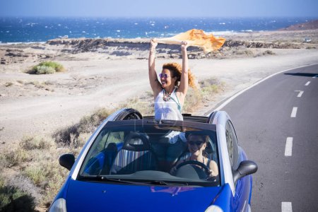 Téléchargez les photos : Voiture Voyage amis transport deux femmes heureuses profiter convertible auto ensemble en vacances voyage d'été - océan et plage en arrière-plan - conduire et avoir un concept amusant - en image libre de droit