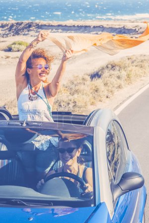 Foto de Viajes en coche amigos transporte dos mujeres felices disfrutan de auto convertible juntos en vacaciones de verano viaje - océano y playa en el fondo - conducción y divertirse concepto - Imagen libre de derechos