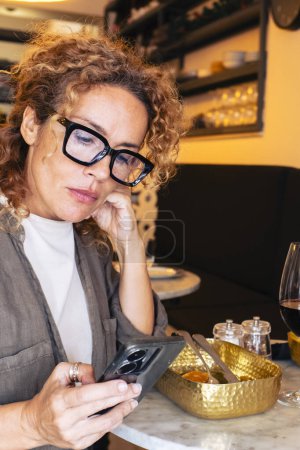 Téléchargez les photos : Femme malheureuse dans un restaurant regardant le téléphone portable - en image libre de droit