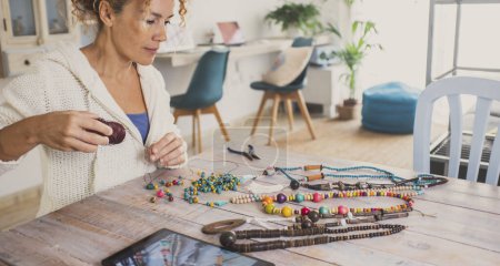 Foto de Busy woman at home start new small business doing jewelry with beads and using laptop to sell on line or learn from tutorial. Indoor hobby leisure activity female people at the table alone - Imagen libre de derechos