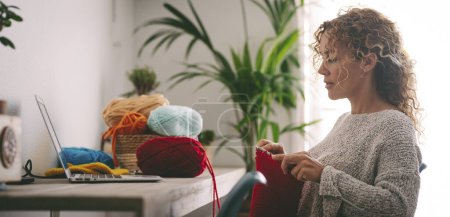 Téléchargez les photos : Femme assise au bureau crochetant une boule de laine rouge suite à un tutoriel sur son ordinateur portable apprendre l'artisanat. - en image libre de droit