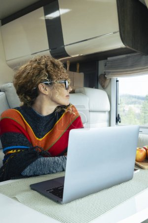 Foto de Vivir y trabajar dentro de la caravana es un estilo de vida digital libre de nómadas. Una mujer sentada en una caravana disfruta de relajarse y conectarse a su computadora portátil. bosque en el fondo fuera de la ventana - Imagen libre de derechos