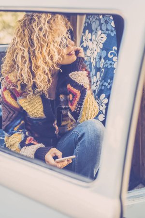Téléchargez les photos : Belle femme aux cheveux bouclés blonds regarde par la fenêtre de son van. printemps, nature et voyage souriant excité par l'aventure et le mode de vie hipster - en image libre de droit