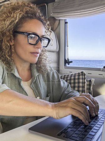 Téléchargez les photos : Femme caucasienne bouclée avec des lunettes de travail sur son ordinateur portable dans son camping-car. vacances à la maison sur roues et aventure de printemps au bord de la mer. Concept de style de vie nomade - en image libre de droit