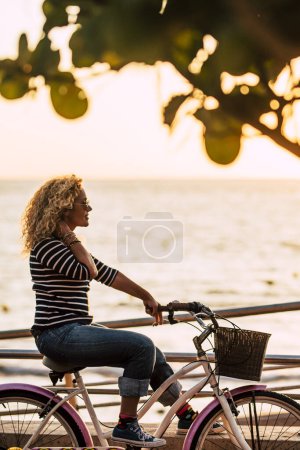 Photo for Outdoor leisure activity for happy people concept with beautiful curly blonde adult woman enjoying the sunset riding her colored bike - active trendy female looking ocean - Royalty Free Image