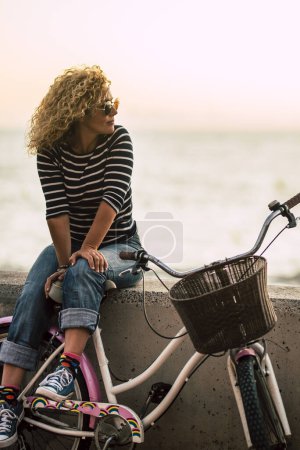 Téléchargez les photos : Les gens profitant d'activités de loisirs en plein air à la recherche d'un beau coucher de soleil coloré - belle jeune femme adulte avec de longs cheveux blonds bouclés profiter de la détente après une visite à vélo - horizon et soleil avec océan - en image libre de droit