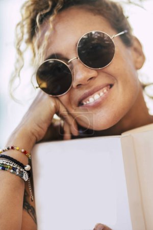 Foto de Alegre hermoso retrato de mujer adulta - señora de mediana edad caucásica con libro de papel blanco y gafas de sol sonrisa a la cámara - enfoque en pulseras accesorios de moda - Imagen libre de derechos