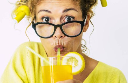Foto de Colores amarillos y sorprendida divertida hermosa mujer adulta caucásica con rizadores en el pelo y fruta naranja para beber estudio y fondo blanco gafas negras y gente bonita - Imagen libre de derechos