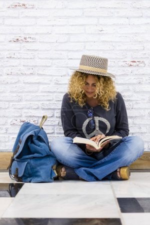 Photo for Alternative trendy travel people lifestyle beautiful curly fashion adult woman sit down on the floor and read a book with her blue backpack near her - white bricks background - Royalty Free Image