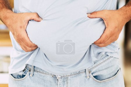 Téléchargez les photos : Homme gras caucasien tenir son ventre avec les mains - régime alimentaire et mode de vie sain concept - les résultats de la malbouffe - la maison en arrière-plan et la quarantaine coronavirus résultat - en image libre de droit