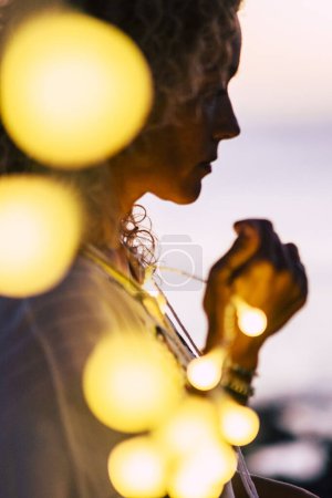 Photo for Emotional concept of hope and faith with adult woman and yellow bulb lights defocused - ocean and sunset in background - beautiful portrait for hoping conceptual image - Royalty Free Image