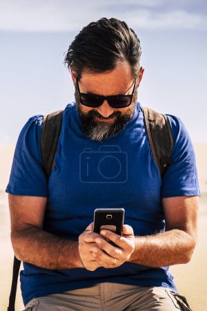Foto de El hombre caucásico adulto usa la conexión a Internet con un teléfono moderno en medio de las dunas del desierto arena - la gente viaja y el concepto de aventura - planeta cálido y peligro de cambio climático - Imagen libre de derechos