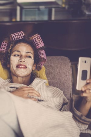 Photo for Cheerful adult woman at home enjoying time with phone video call while have a beauty mask on face and curlers on hair for beauty skin and hairstyle activity - Royalty Free Image
