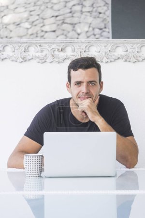 Photo for Young business manager handsome man with blue eyes sitting on the desktop with computer laptop looking at the camera - luxury lifestyle for caucasian people at home - Royalty Free Image