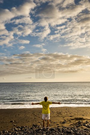 Téléchargez les photos : Coucher de soleil coloré sur la plage et jeune homme debout et profitant de la beauté de la nature et de l'océan - grand ciel et horizon en arrière-plan - en image libre de droit