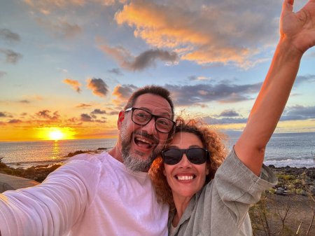 Téléchargez les photos : Couple heureux et joyeux d'homme et de femme adultes prenant ensemble une photo selfie avec téléphone portable à la plage contre un magnifique coucher de soleil de couleur sur l'océan. Voyage aventure touriste s'amuser en vacances - en image libre de droit